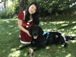 Eine Person in einem roten Kleid mit weißen Knöpfen kniet neben einem schwarz-weißen Hund an der Leine, der auf dem Gras liegt. Im Hintergrund sind Bäume zu sehen.