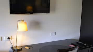 A hotel room with a flat-screen TV mounted on the wall, a desk with a lamp, a chair, and a remote control on the desk.