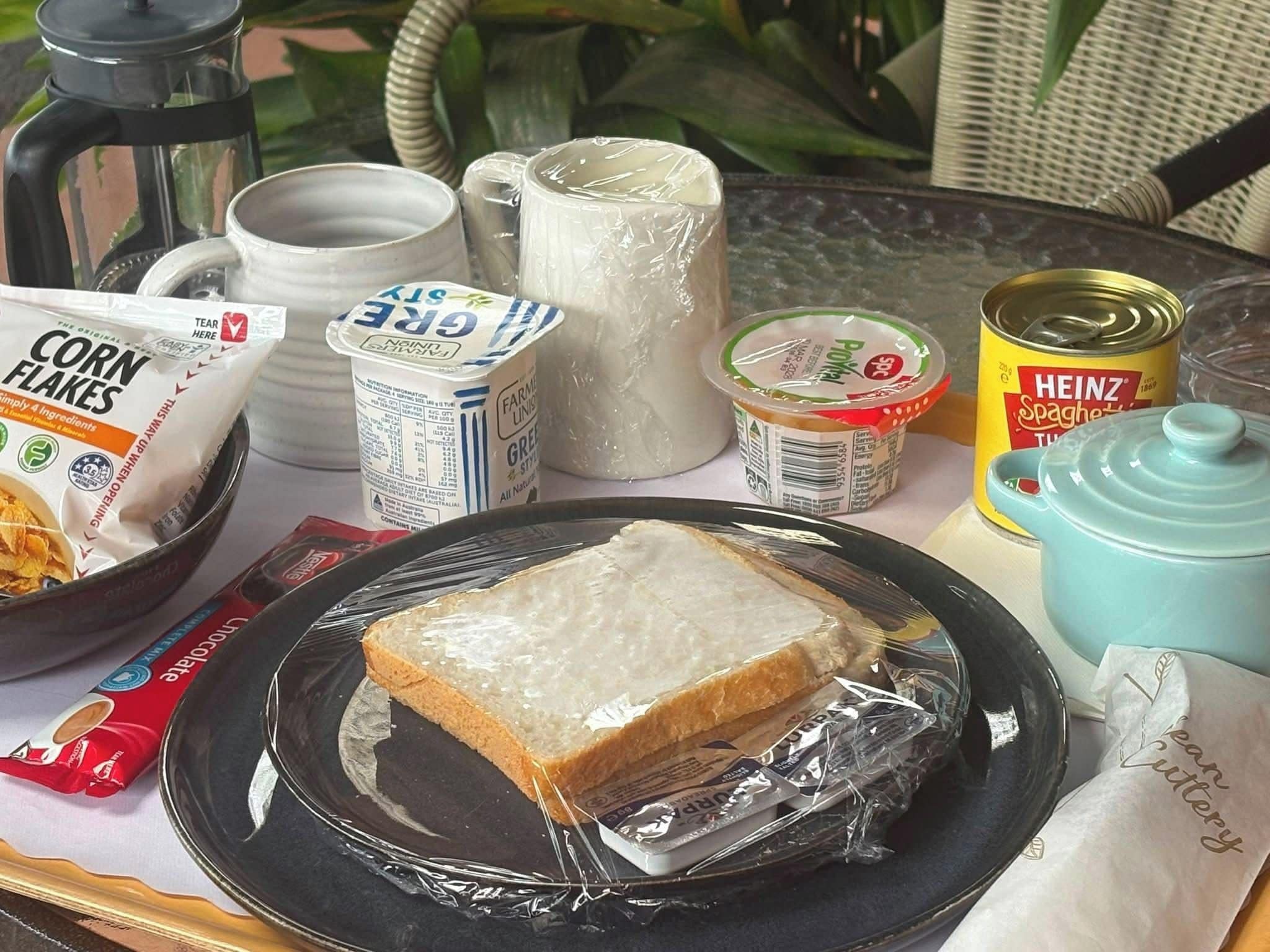 Una mesa con un plato de pan, una lata de Spaghetti Heinz, un tarro de copos de maíz, una jarra de leche, un termo y una tetera.