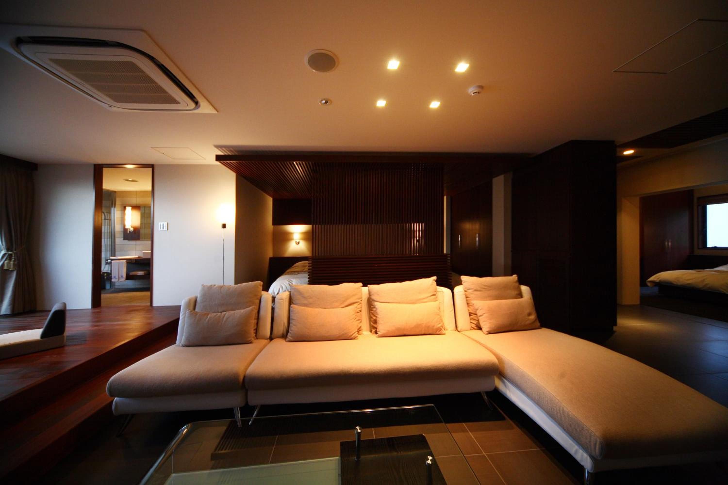 A modern living room with a sectional sofa, a glass coffee table, and a view of the adjacent bedroom.