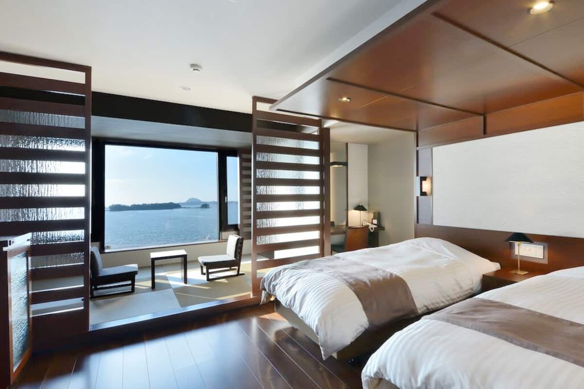 A modern hotel room with two beds, a seating area, a large window with a view of the sea, and a wooden partition.