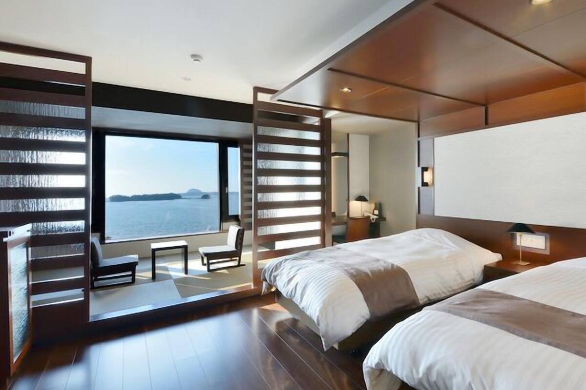A modern hotel room with two beds, a large window with a view of the sea, a seating area with a table and chairs, and a wooden partition.
