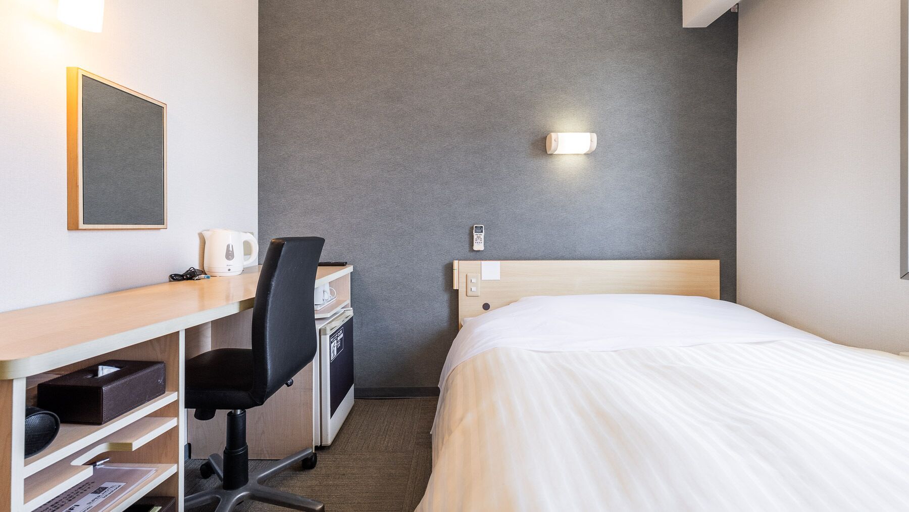 A hotel room with a bed, desk, chair, and a small table with a kettle.