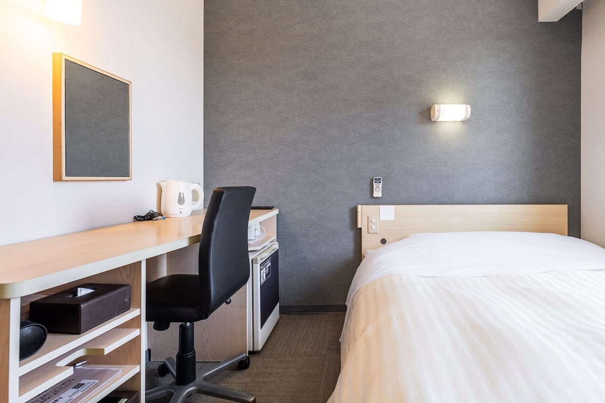 A hotel room with a bed, desk, chair, and a small table with a kettle.