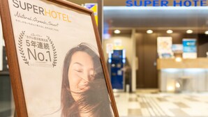 A framed sign with text and a blurred photo, placed in a hotel lobby with a reception desk and a blue sign reading "SUPER HOTEL". - Super Hotel Fuji Inter (Fuji)