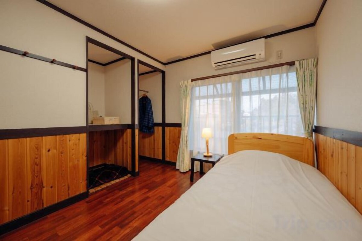 Un dormitorio con piso de madera, una cama con colchón blanco, una mesita de noche con lámpara, una ventana con cortinas y un equipo de aire acondicionado.