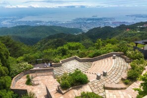 Un anfiteatro en terrazas con escalones de piedra y una zona de asientos, rodeado de exuberante vegetación y con vistas a una ciudad costera.