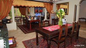 A dining area with a wooden table, chairs, and a chandelier. There are paintings on the walls and a window with curtains.