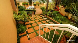 A paved garden pathway leading to a car parked in a driveway, surrounded by lush greenery and a white railing.