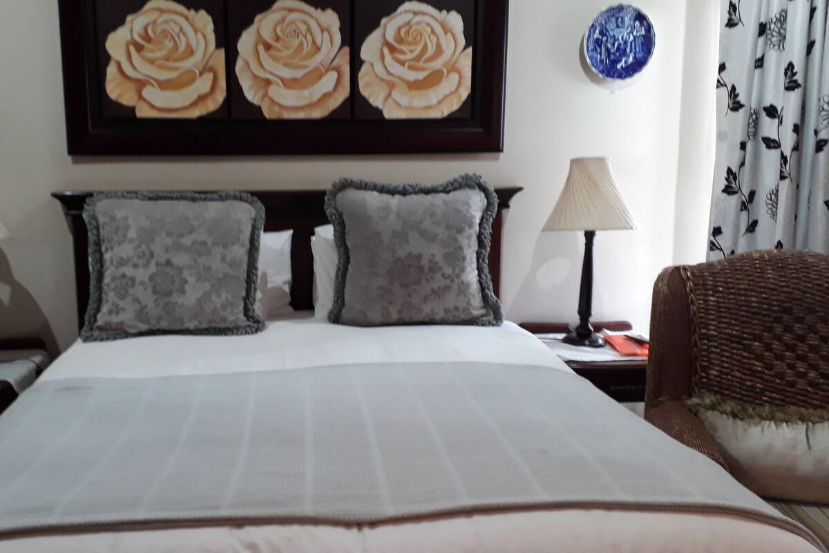 A neatly made bed with two pillows, a nightstand with a lamp, and a wall with a framed picture of roses.