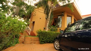 A two-story building with a staircase leading to the entrance, surrounded by greenery and a parked car.