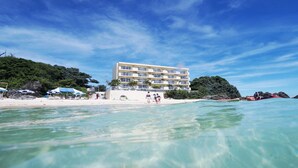 Un hotel frente a la playa con vista despejada al océano, una playa de arena y abundante vegetación.