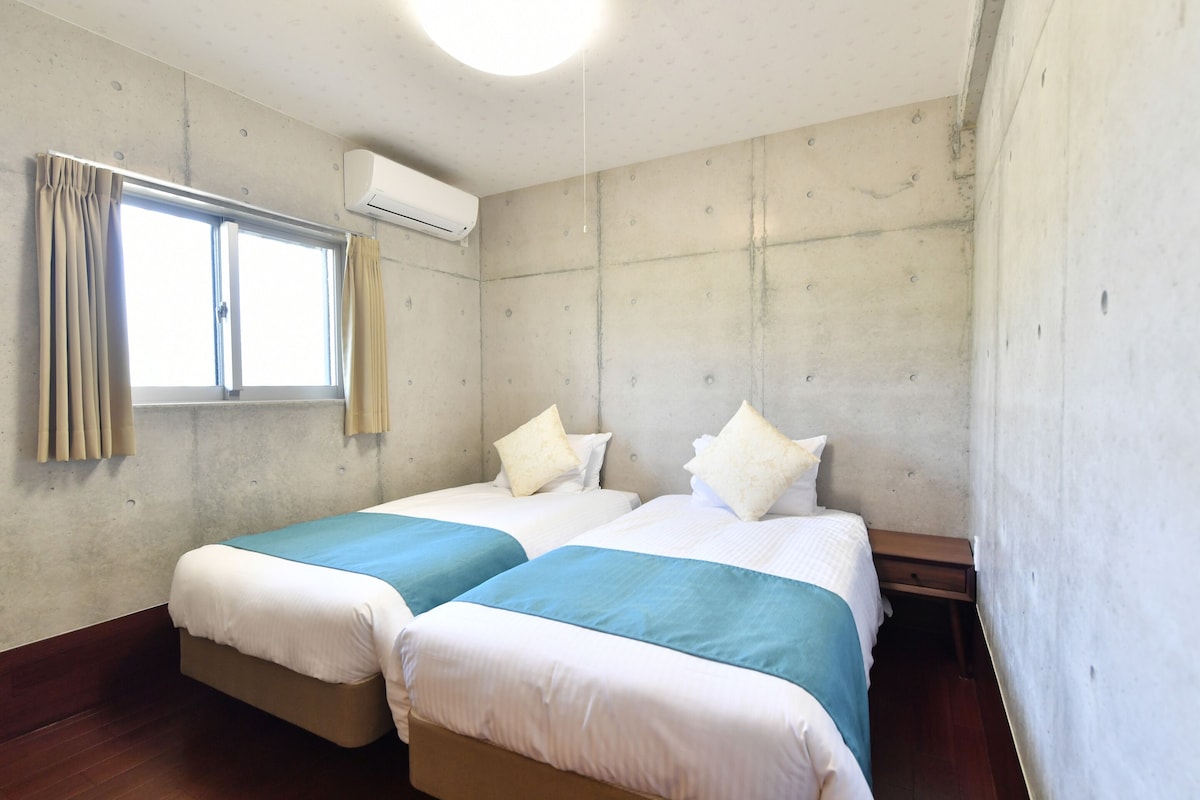 A hotel room with two beds, a window with curtains, an air conditioning unit, and a wooden floor.