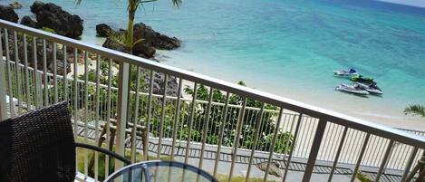 A balcony with a glass table and wicker chairs overlooking a beach with clear turquoise water and a few boats.