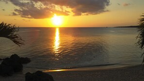 A sunset over a calm sea with rocks in the foreground and a distant landmass. - Churaumi On The Beach Motobu (Motobu)