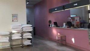 A hotel reception area with a pink counter, a clock, and a sign indicating the front desk. There are stacks of white pillows on a cart and a small stool near the counter. - Hotel Crown Hills Kofu (Kofu)