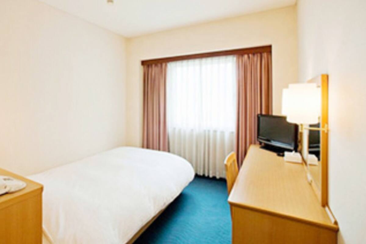 A hotel room with a single bed, a desk, a television, and a window with curtains.