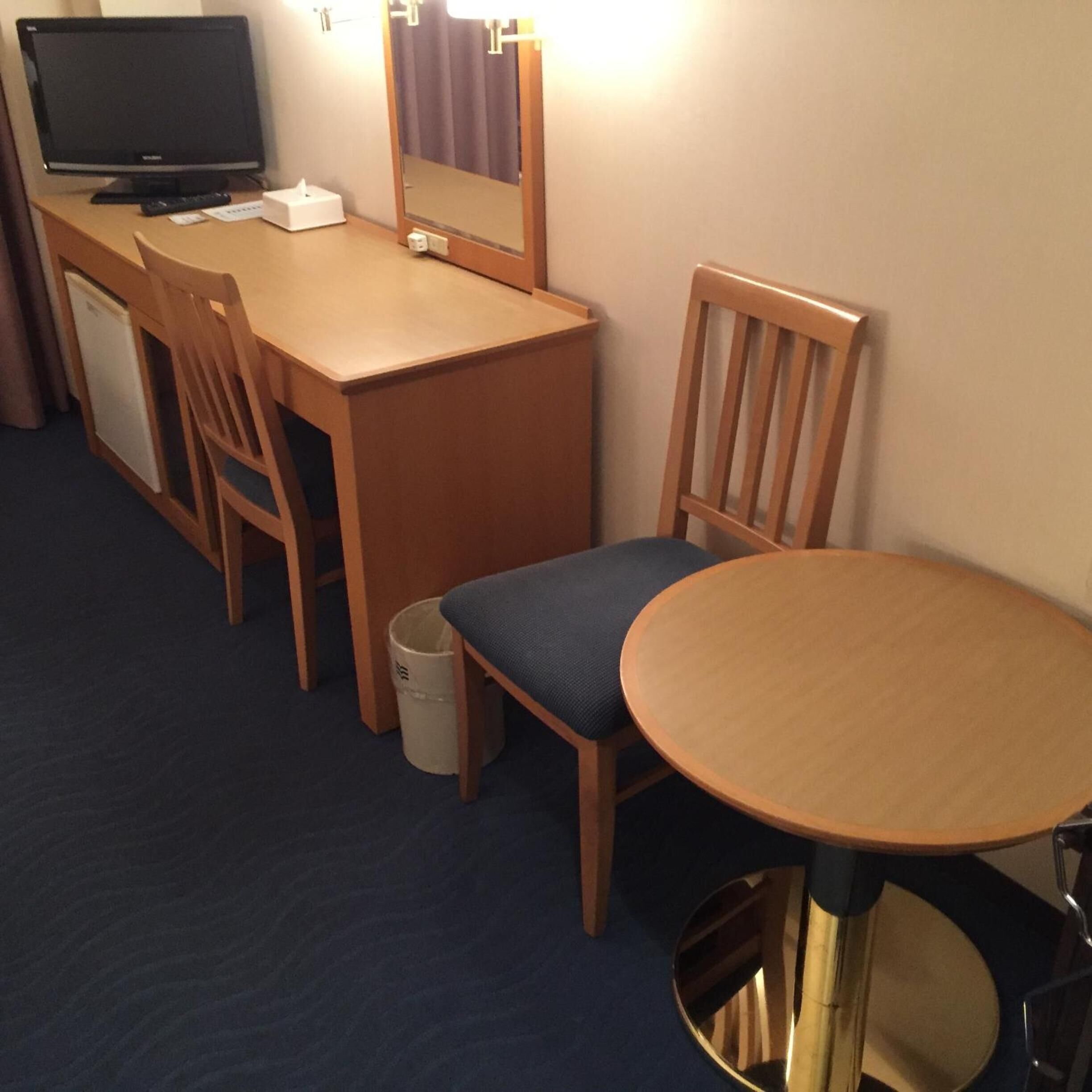 A hotel room with a wooden desk, a television, a mirror, a chair, and a small round table.