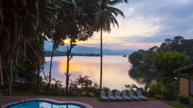 A swimming pool with lounge chairs and a view of a calm lake at sunset. - Oruthota Chalets (Maberiyatenna)