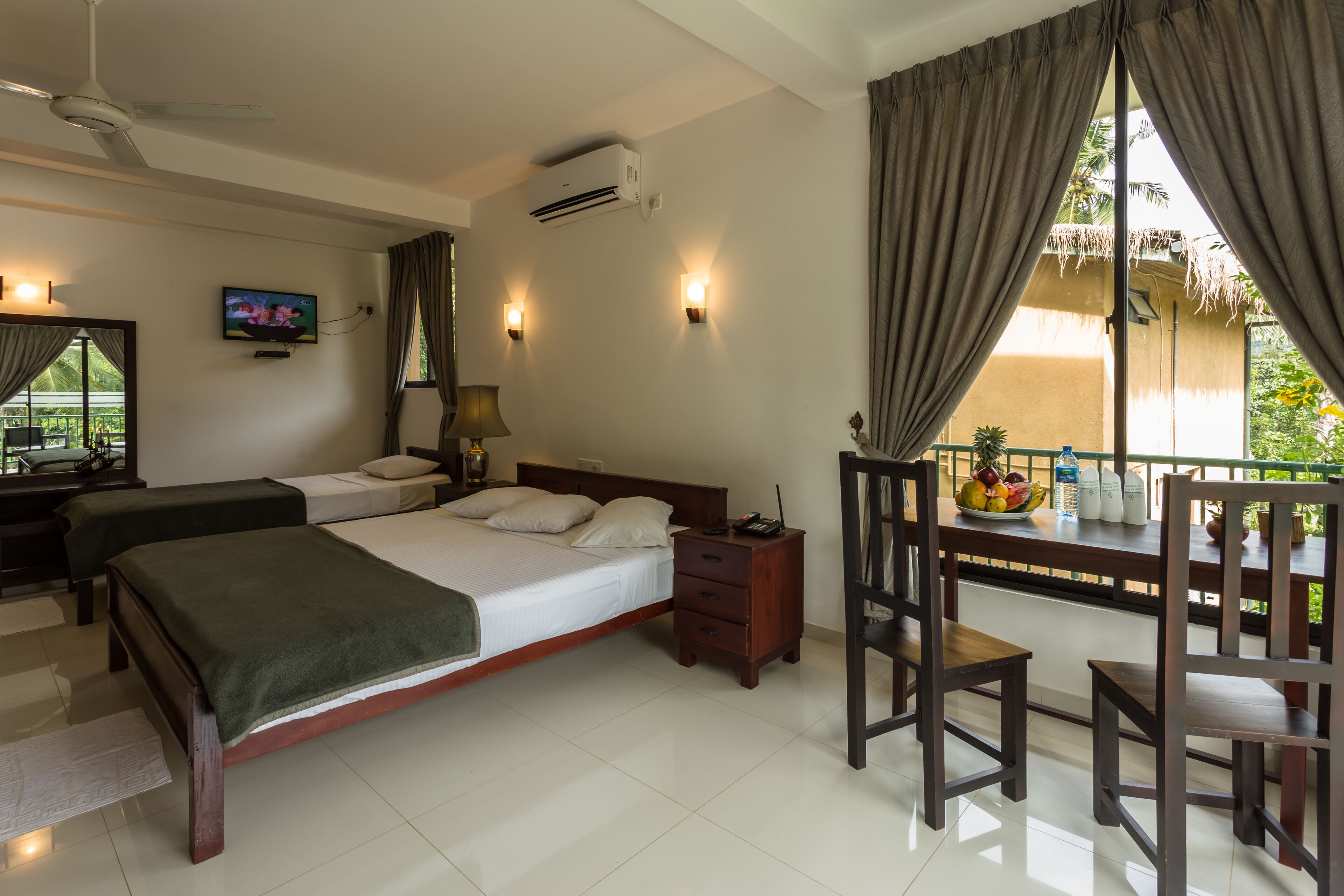 A hotel room with two beds, a TV, a desk with a chair, a balcony with a table and chairs, and a view of greenery.
