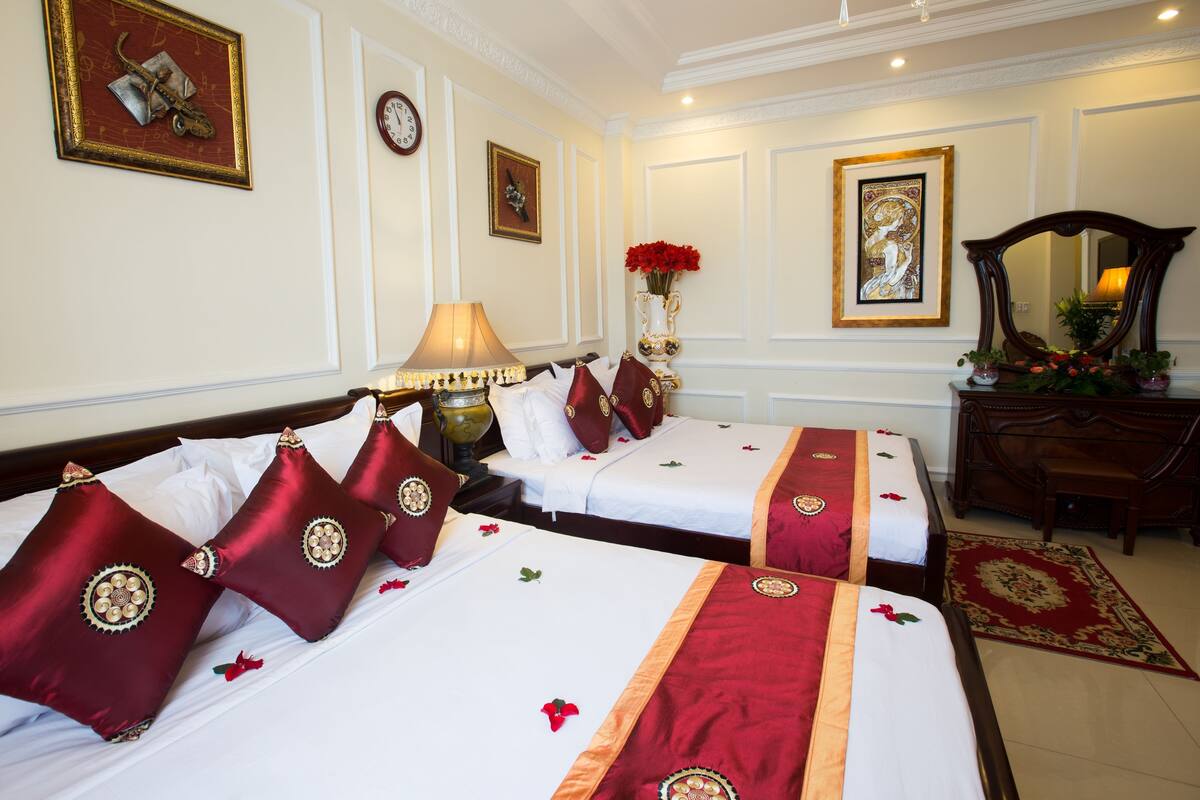 A hotel room with two beds, a nightstand, a lamp, a vase with flowers, a clock, and a mirror.