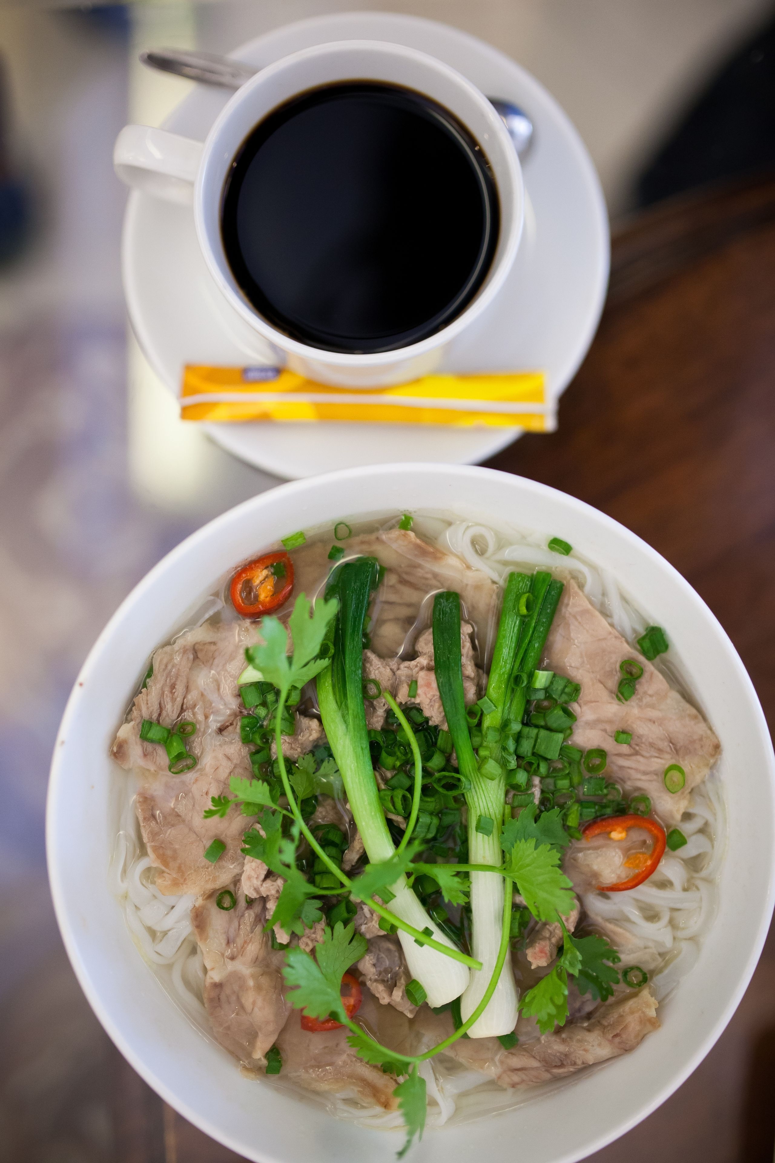 A bowl of noodle soup with meat and vegetables, accompanied by a cup of black coffee.