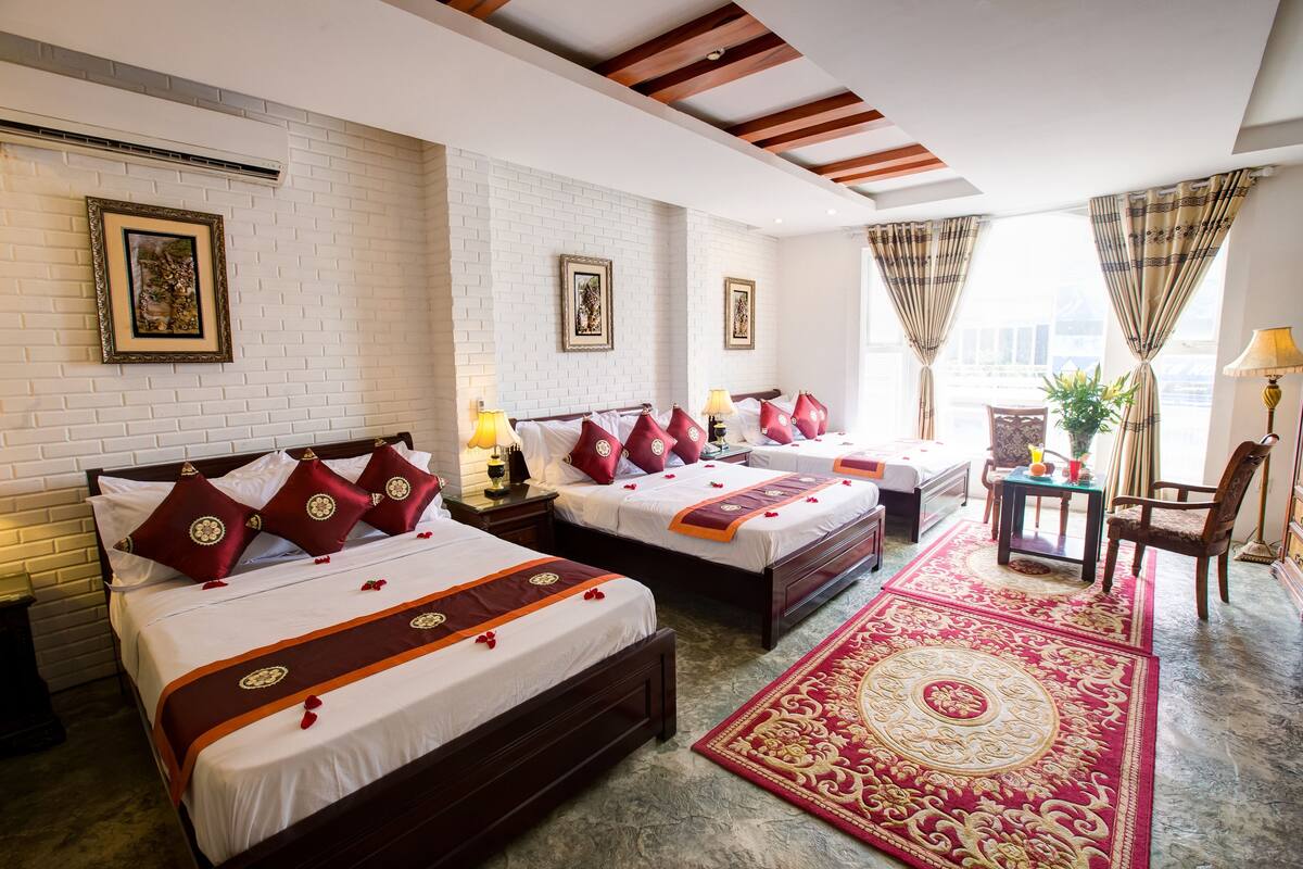 A hotel room with three beds, each with red and white bedding, a red and gold patterned rug, and framed artwork on the walls.