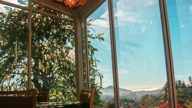 Un invernadero con una mesa y sillas, ventanas amplias y vistas a una montaña y flores rojas.