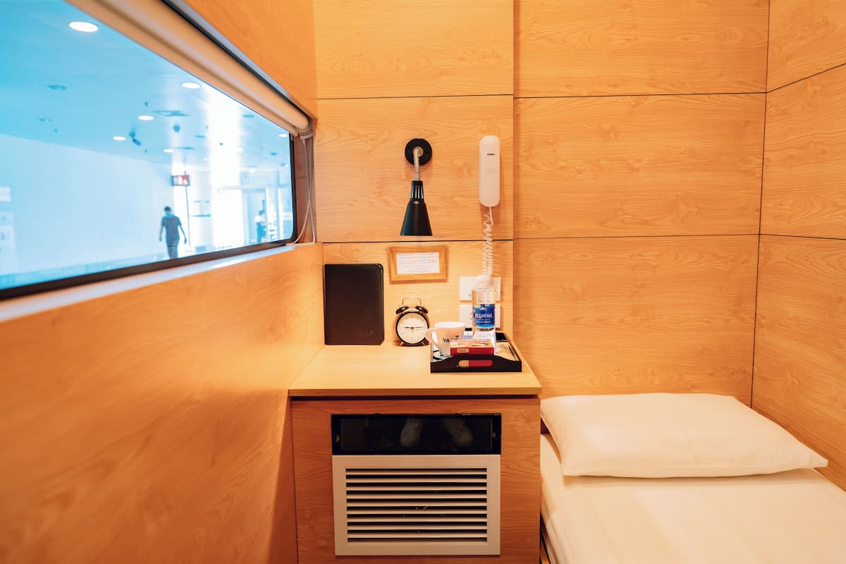Habitación de hotel con pared de paneles de madera, mesita de noche con lámpara, teléfono, cuadro enmarco, reloj pequeño y ventana con vistas al exterior.