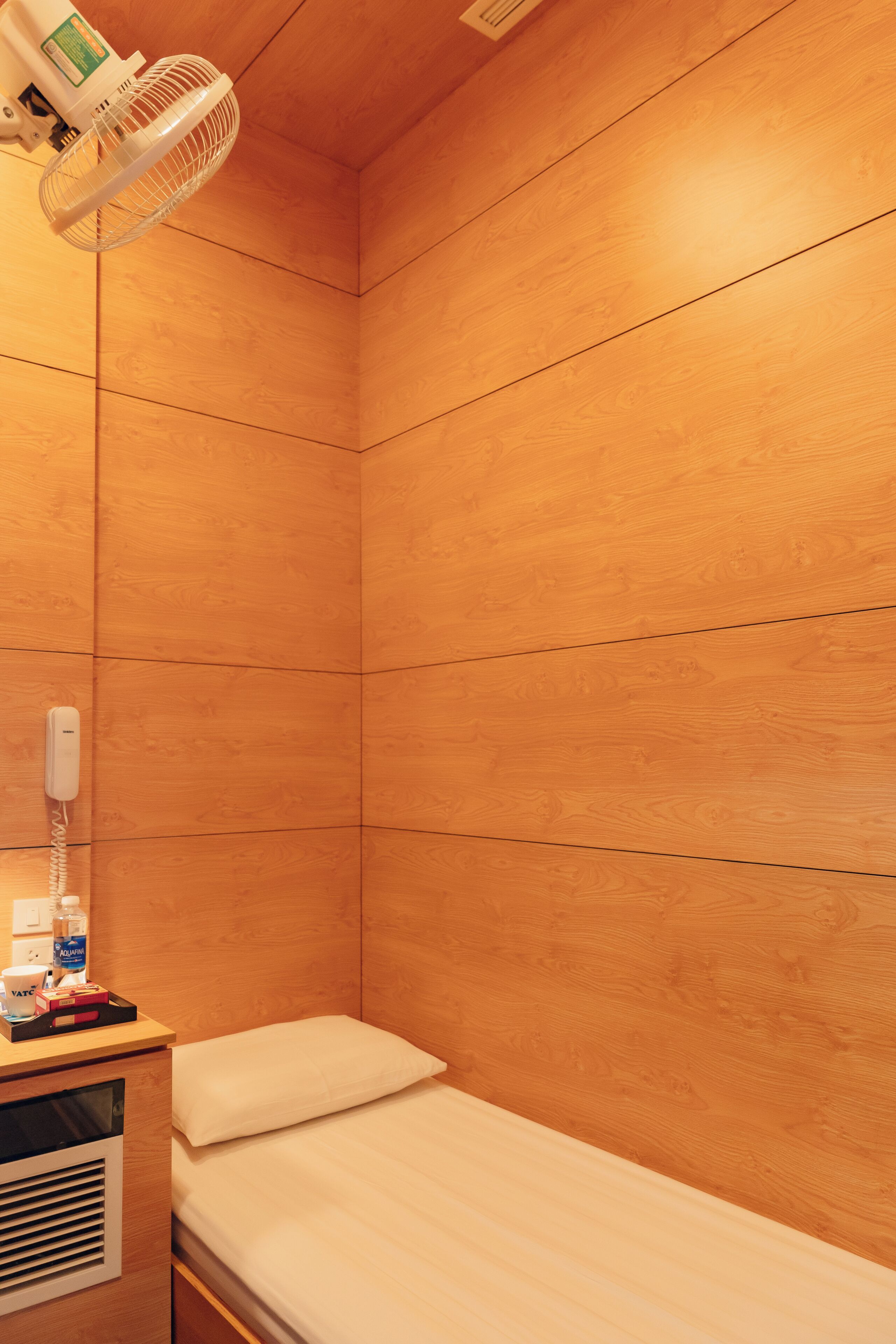 Una habitación pequeña con cama individual, pared de madera, mesita de noche con lámpara, ventilador y teléfono.