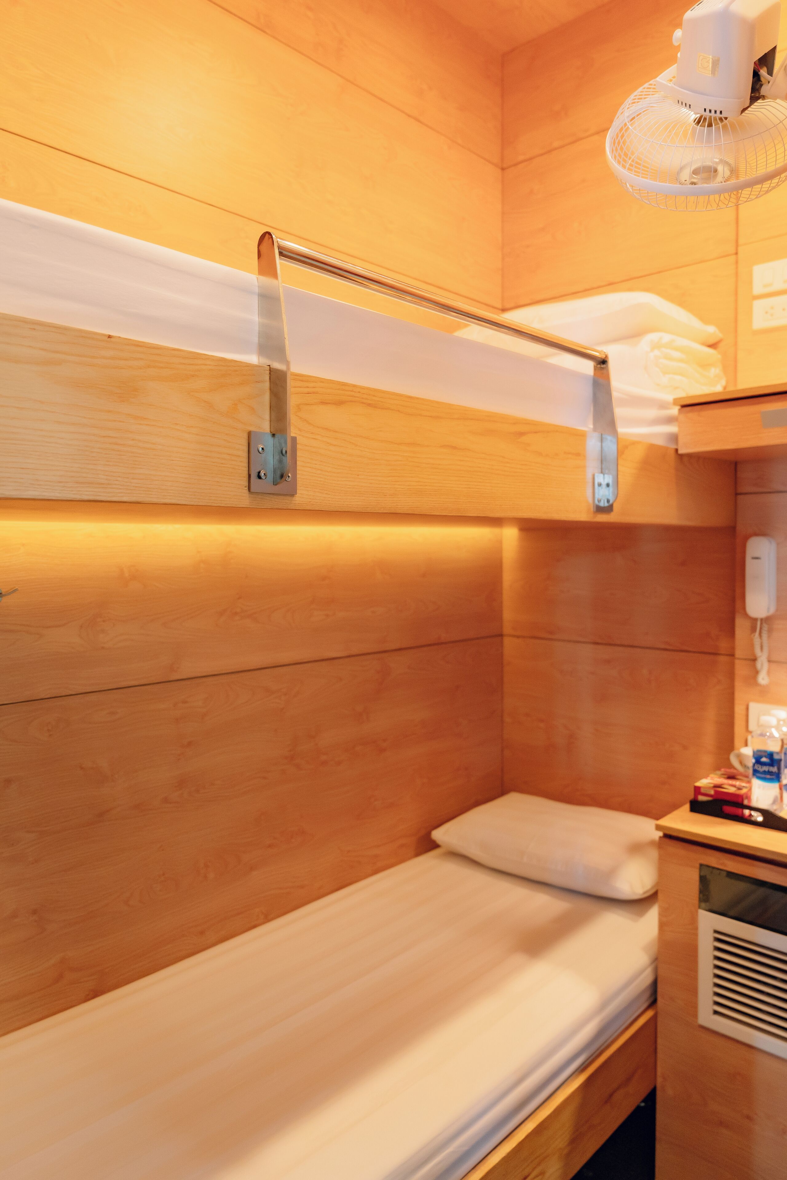 Una cama individual con cabecera, una mesita de noche con lámpara y un ventilador de pared.