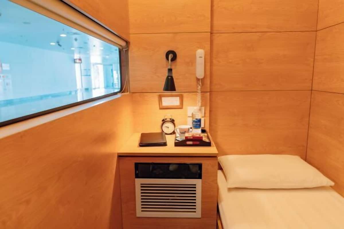 Una habitación pequeña y funcional con ventana, escritorio con lámpara, teléfono, reloj y cama.