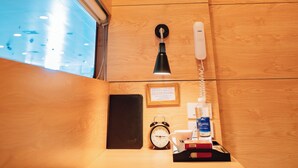 Une chambre d’hôtel avec un plancher en bois, un bureau avec un téléphone, une petite horloge, un ventilateur et une fenêtre donnant sur le ciel.