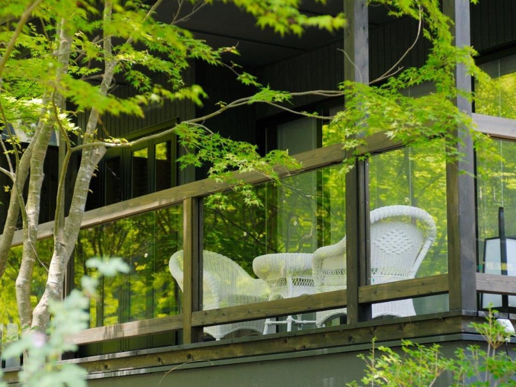 A wooden balcony with a glass enclosure, white wicker furniture, and a view of trees.