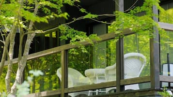A wooden balcony with a glass enclosure, white wicker furniture, and a view of trees.