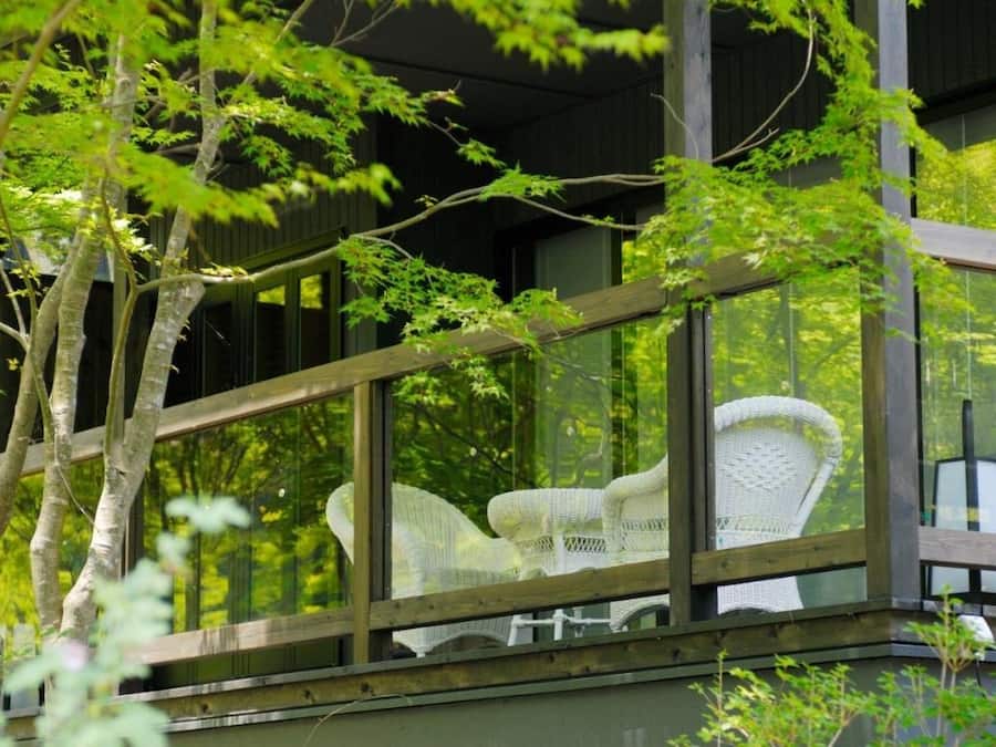A wooden balcony with a glass enclosure, white wicker furniture, and a view of trees.