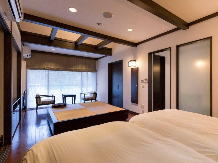 Een hotelkamer met een bed, een bureau en een stoel. Er is een raam met gordijnen, een deur en een wandlamp.