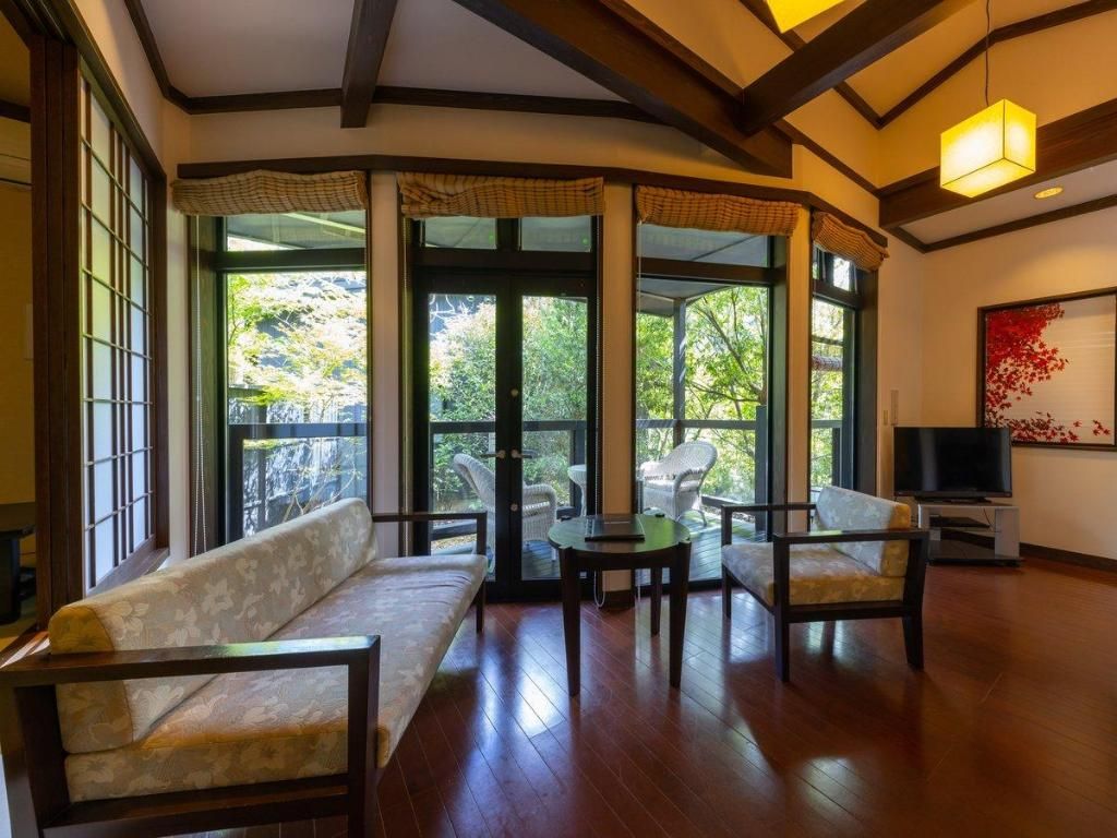 A room with a wooden floor, a sofa, a table, and chairs. There are large windows with a view of trees.