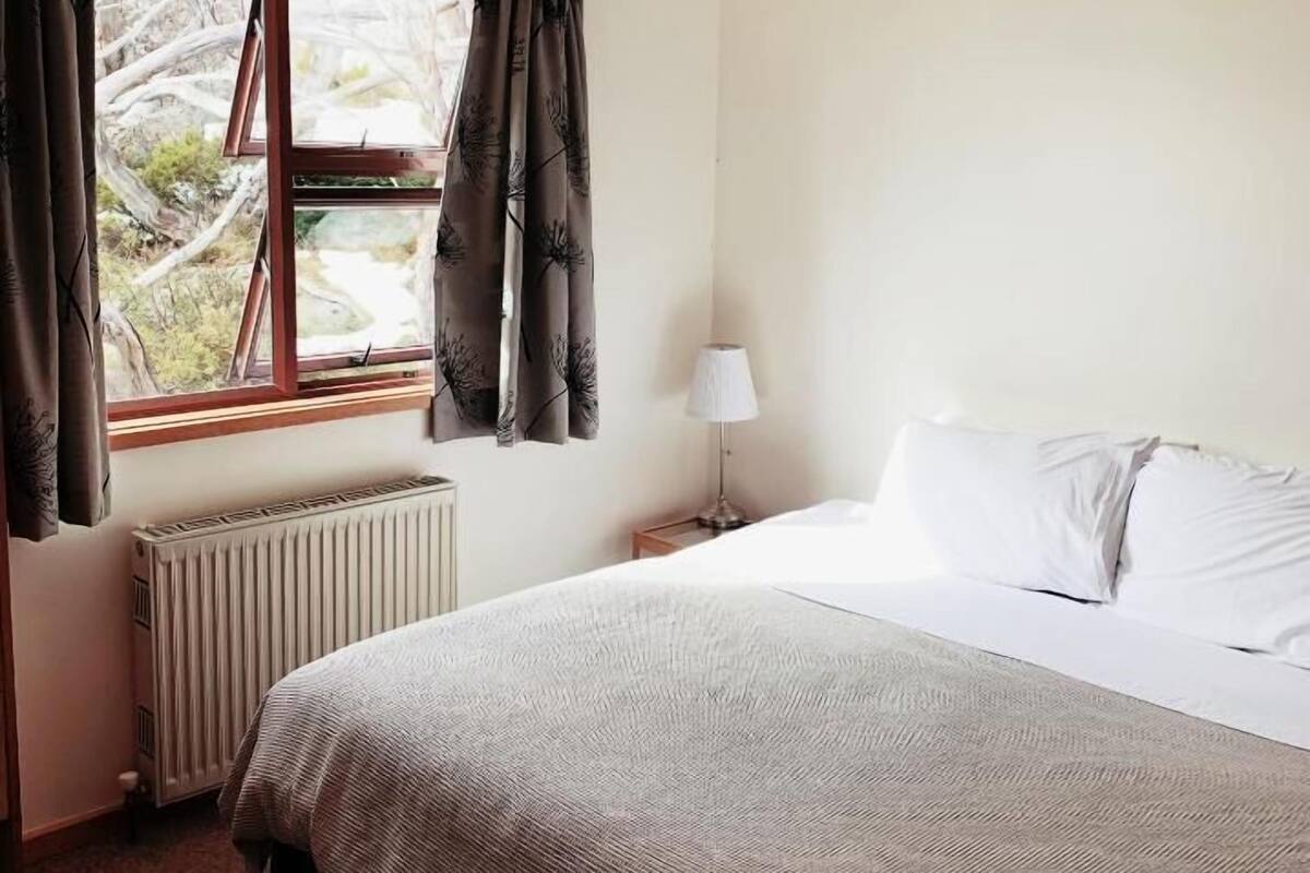 Un dormitorio con cama, ventana con cortinas, radiador y mesita de noche con lámpara.