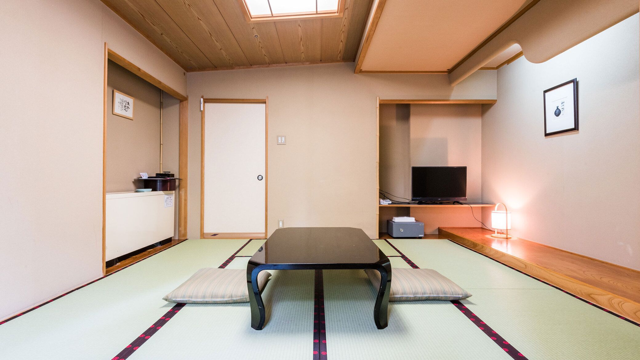 Une pièce traditionnelle avec un sol recouvert de tatamis, une petite table basse, des coussins, un téléviseur et une petite étagère où sont posés divers objets.