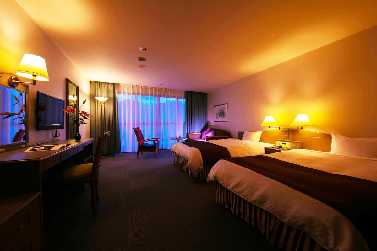 A hotel room with two beds, a desk, a chair, a TV, and a window with curtains.