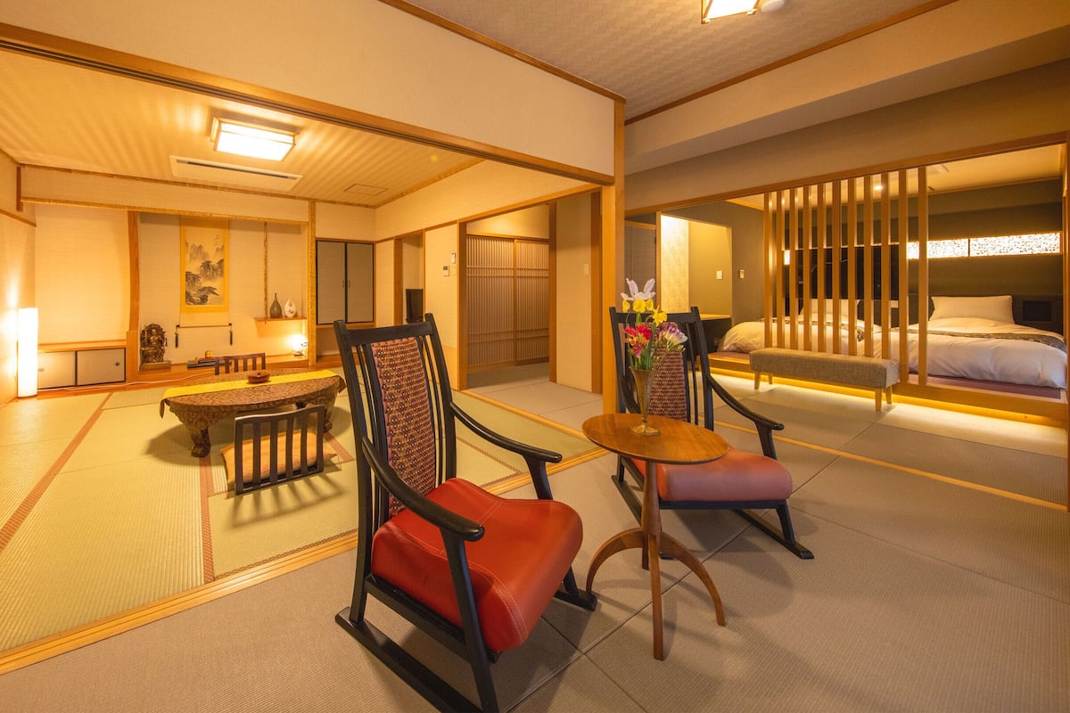 A traditional Japanese-style hotel room with tatami flooring, a low table, two chairs, a bed with a wooden headboard, and a sliding shoji screen.