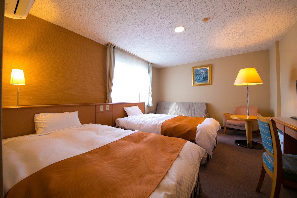 A hotel room with two beds, a desk, a chair, and a lamp.