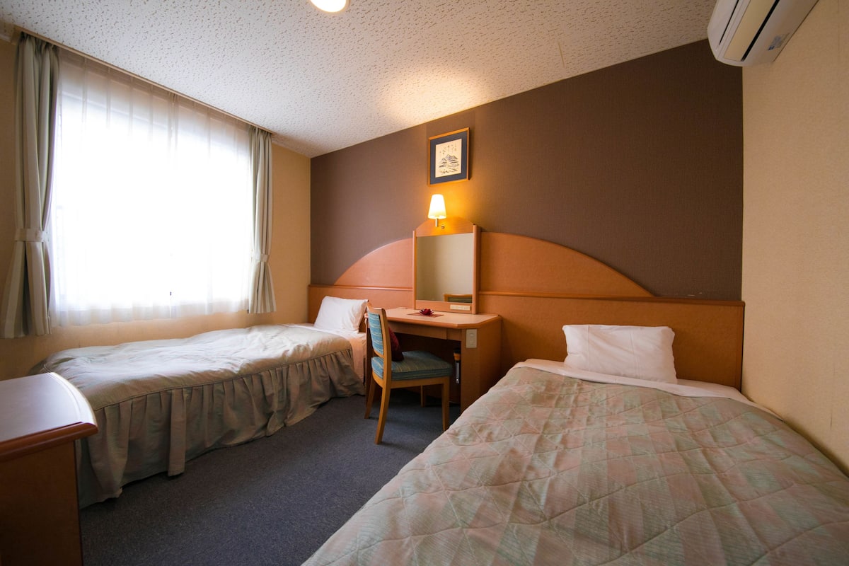 A hotel room with two single beds, a desk with a chair, a lamp, and a framed picture on the wall.