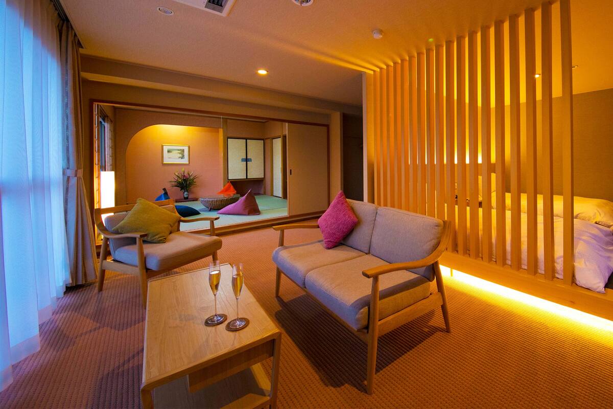 A hotel room with a sofa, a small table with glasses, a bed with pillows, a sliding door, and a view of the outside.