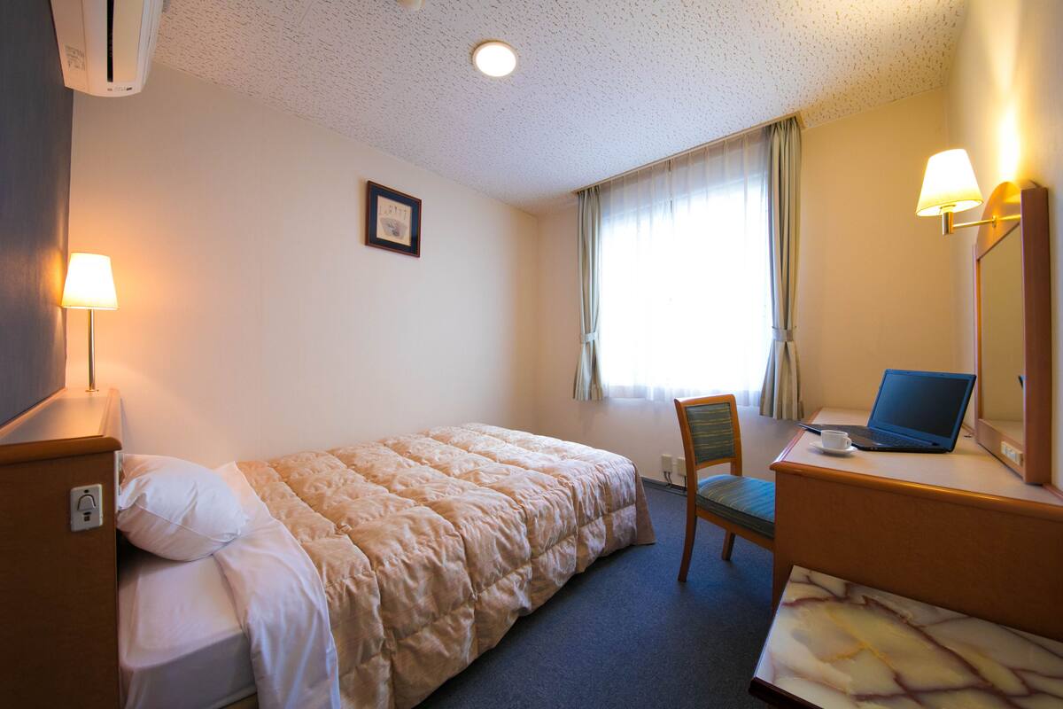 A hotel room with a bed, a desk with a laptop, a chair, and a lamp.