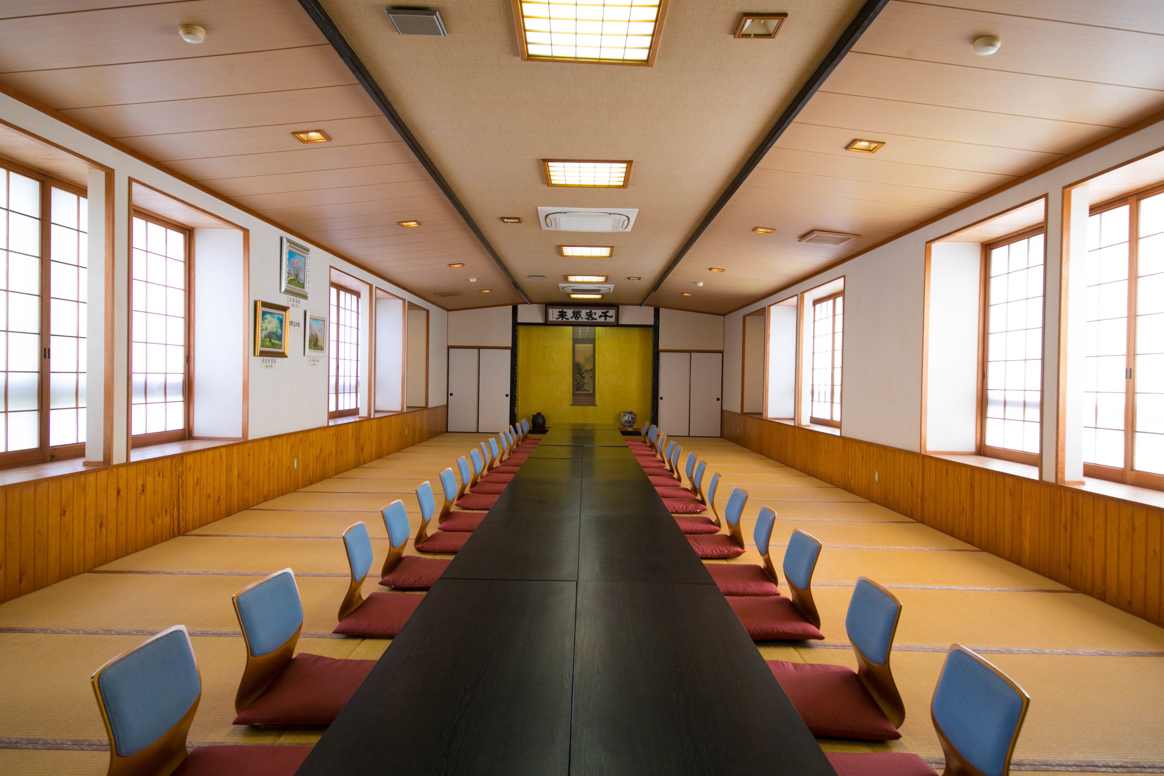 A long room with rows of tatami mats and blue cushions, wooden walls, and large windows.