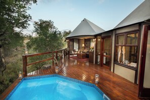 A wooden deck with a swimming pool, a lounge area with a sofa and table, and a large window.