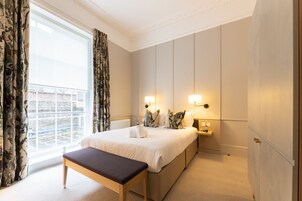 Quarto de hotel com uma cama grande, um banquinho, uma mesa de cabeceira e vista da cidade através de uma janela com cortinas.