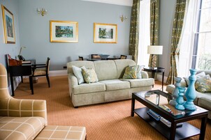 A living room with a sofa, armchair, coffee table, and a dining area with a table and chairs. There are framed pictures on the wall and a lamp on a side table.